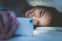 Girl lying on bed under pillow and using smartphone — Stock Photo