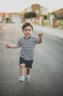 Retrato de lindo niño en pantalones cortos y camiseta corriendo felizmente hacia la cámara en la carretera . - foto de stock