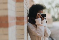Вид збоку дівчини, спираючись на цегляну стіну і роблячи фотографії з аналоговою камерою — стокове фото