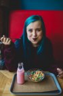 Portrait de fille aux cheveux bleus assise à une table de café, mangeant des céréales avec une cuillère et regardant la caméra — Photo de stock