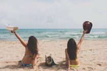 Rear view of two girlfriends in bikini sitting on beach with hats in raised arms — Stock Photo
