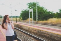 Portrait of young girl with red hair talking over smartphone on railway countryside platform — Stock Photo