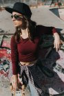 Молодая стильная девушка в шляпе и солнцезащитных очках позирует со скейтбордом под рукой и смотрит в скейтпарк. — стоковое фото