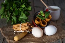 Still life of fried honey dough tubes with mint in mug lying on sacking with eggs over rural wooden table — Stock Photo