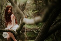 Спокойная девушка в белом платье сидит на ветке в лесу — стоковое фото