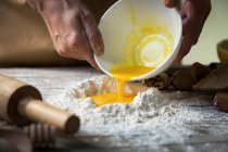 Vue rapprochée des mains mettant des œufs écrasés dans un tas de farine sur une table de cuisine en bois — Photo de stock