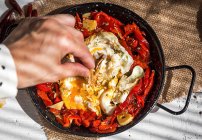 Vista superior de la mano poner ingrediente en la sartén con huevos revueltos y tomates secos - foto de stock