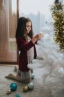 Side view of adorable girl sitting on floor and putting baubles on white decorative Christmas tree — Stock Photo