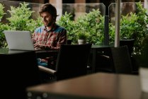 Portrait of bearded man in checkered shirt sitting at cafe terrace table and working on laptop — Stock Photo