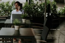 Vista à distância do empresário sorridente sentado à mesa e falando sobre smartphone enquanto usa laptop no terraço do café — Fotografia de Stock