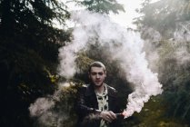 Retrato de hombre con estilo agitando la mano con vela de humo entre los bosques y mirando a la cámara - foto de stock
