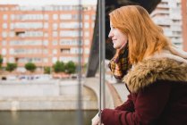 Vista laterale della donna dai capelli rossi che distoglie lo sguardo e sorride in piedi sul ponte — Foto stock