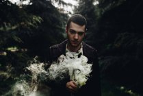Портрет стильного человека с дымовой свечой, позирующей в лесу и смотрящего в камеру — стоковое фото