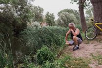 Vue latérale de l'homme âgé accroupi sur la rive du lac avec vélo appuyé sur l'arbre — Photo de stock