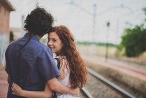 Вид сзади девушки, смотрящей через плечо в камеру, обнимающейся с парнем на железнодорожной платформе — стоковое фото