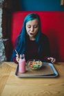 Портрет синьоволохатої дівчини-підлітка, що сидить за столом у кафе і дивиться вниз на піднос з йогуртом і мискою з різнокольорових зернових — стокове фото