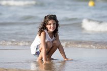 Fille bouclée assis à la plage et regardant la caméra — Photo de stock