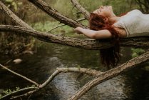 Ginger girl lying on bending over river tree branch — Stock Photo
