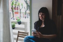 Жінка сидить біля вікна і використовує якрозумний телефон — стокове фото