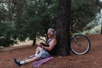 Seitenansicht eines älteren Mannes, der mit Buch in der Hand auf dem Boden sitzt und sich mit einem geparkten Fahrrad an einen Baum lehnt — Stockfoto