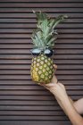 Female hand holding funny pineapple with sunglasses over brown shutters — Stock Photo