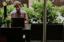 Вид спереду усміхненого чоловіка в картатій сорочці, що сидить за столом тераси кафе з горщиком і використовує ноутбук — стокове фото