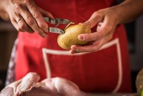 Primer plano de manos femeninas pelando patata cruda para asar con pollo - foto de stock