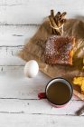 Плоский вид на лимонный торт на хлебобулочной бумаге и кружку горячего шоколада на белом сельском столе с яйцом — стоковое фото
