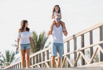 Lächelnde Familie mit Tochter, die auf den Schultern des Vaters sitzt und im Sonnenlicht die Brücke entlangläuft. — Stockfoto