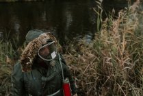Vue grand angle de l'homme portant manteau avec capuche et masque à gaz debout sur la rive du lac et regardant loin — Photo de stock