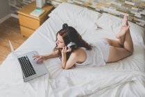 Mujer en la cama usando el portátil y escuchando música - foto de stock