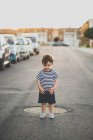 Niño posando en la cámara mientras está de pie en el camino de asfalto en el suburbio - foto de stock