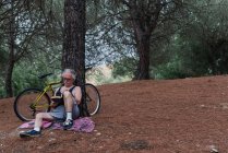 Vista frontal do homem sênior sentado à árvore e lendo o livro ao lado da bicicleta estacionada na floresta — Fotografia de Stock