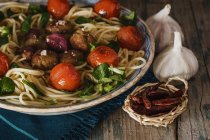Vista de perto de espaguete com almôndegas, tomates cereja e folhas de manjericão em mesa de madeira com pequena cesta de pimentas vermelhas e alho — Fotografia de Stock