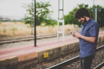 Вид збоку усміхненого чоловіка, що використовує смартфон на залізничній сільській платформі — стокове фото