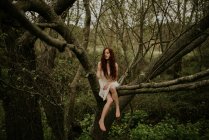 Ginger girl posing on bending over ground tree branch and looking away — Stock Photo