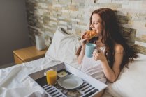 Mädchen isst Croissant mit Tasse Kaffee im Bett — Stockfoto