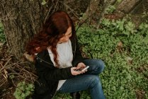 Улыбающаяся девочка сидит у дерева и просматривает смартфон — стоковое фото
