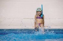 Enfant en masque de plongée assis au bord de la piscine et éclaboussant l'eau avec les jambes — Photo de stock