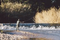 Distant view of man standing on river shore and fishing with rod — Stock Photo