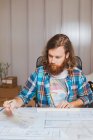 Portrait de l'homme barbu en chemise à carreaux assis sur le lieu de travail et avec des plans au bureau. — Photo de stock