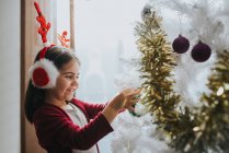 Vista lateral de la chica feliz con orejeras de piel con astas colocando bolas en el árbol de Navidad decorativo - foto de stock