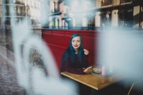Retrato de menina com cabelo azul sentado na mesa de café com smartphone e olhando para longe — Fotografia de Stock