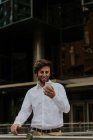 Портрет улыбающегося бизнесмена в белой рубашке, смотрящего на смартфон в руке на городской сцене — стоковое фото