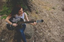 Retrato de alto ângulo de menina de cabelos vermelhos sorridente sentado no rock e tocando guitarra — Fotografia de Stock