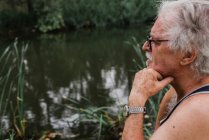 Vista lateral do homem sênior em óculos olhando para longe com a mão no queixo contra o rio do campo no fundo — Fotografia de Stock