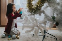 Side view of adorable girl sitting on knees and placing baubles on white decorative Christmas tree in room. — Stock Photo