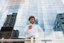 Retrato de baixo ângulo de homem de negócios sorridente em camisa branca usando telefone sobre fachada edifício de negócios — Fotografia de Stock