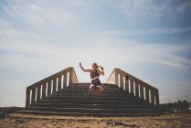 Chica joven en vestido colorido y gafas de sol saltando en la playa contra escaleras de piedra - foto de stock