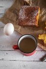 Vista de ángulo alto de taza roja cerca de rodajas caseras de pastel de limón en papel de panadería y huevo en mesa de madera rural - foto de stock